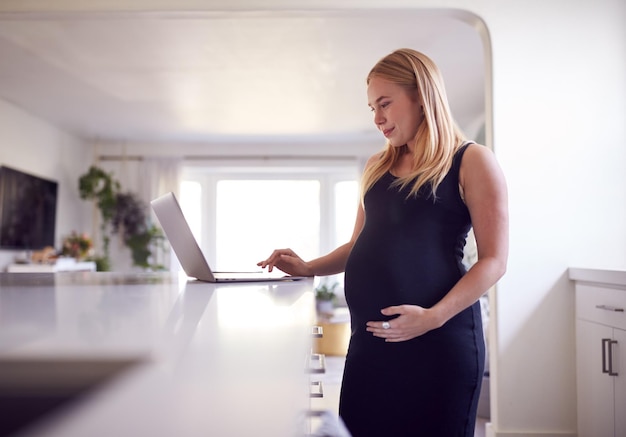 Femme enceinte debout près du comptoir de la cuisine travaillant à domicile sur un ordinateur portable