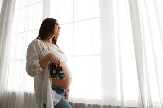 Une femme enceinte debout près de la fenêtre tient les chaussures de l'enfant, caresse le ventre et l'estomac.