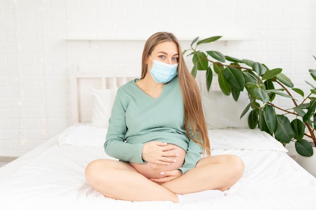 Une femme enceinte est assise dans un masque de protection médicale sur le lit et touche son gros ventre à la maison le concept de grossesse et d'attente de la naissance d'un bébé et d'un long terme