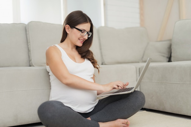 Femme enceinte est assise en tailleur et travaille sur un ordinateur portable gris.