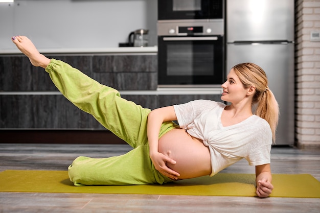 Femme enceinte flexible avec le ventre nu soulevant une jambe en faisant des exercices sur le sol
