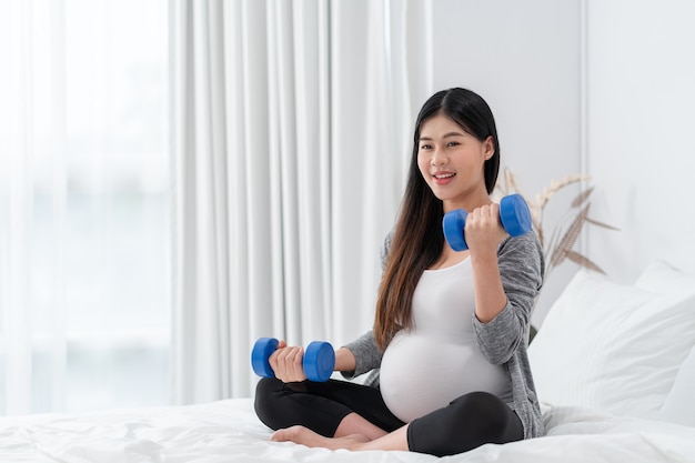 Une femme enceinte heureuse asiatique est assise et fait de l'exercice sur le lit. concept de grossesse, de maternité, de personnes et d'attente