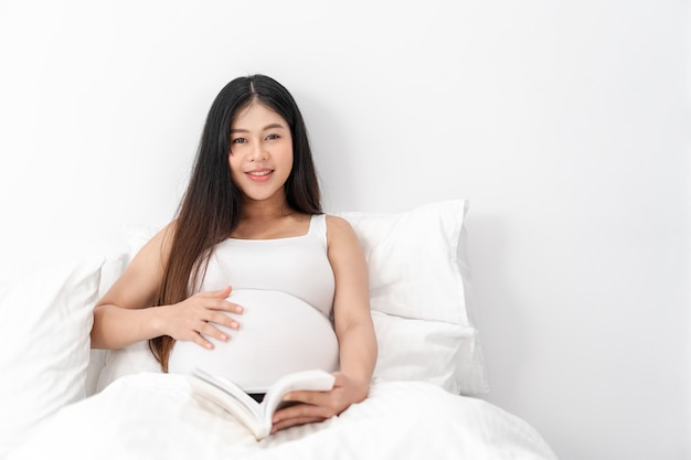 Une femme enceinte heureuse asiatique est assise et lit un livre sur le lit et touche son ventre. concept de grossesse, de maternité, de personnes et d'attente