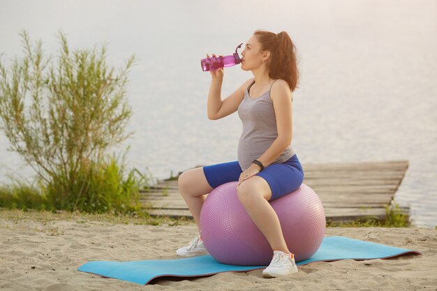 Une femme enceinte heureuse boit de l'eau sur un ballon de gym à l'extérieur