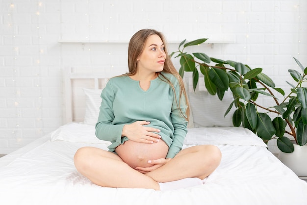 Une femme enceinte heureuse est assise sur le lit et touche son gros ventre à la maison, le concept de grossesse et attend la naissance d'un bébé et un long terme