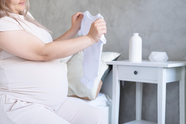 femme enceinte heureuse avec gros ventre tenant des vêtements de bébé dans les mains à la maison. notion de maternité