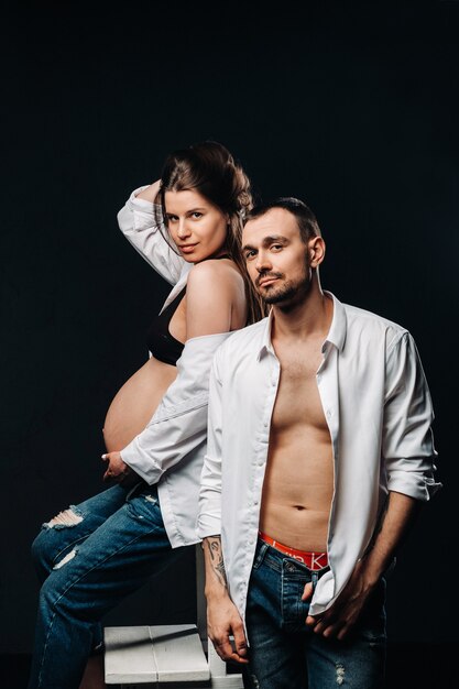 Une femme enceinte et un homme en chemise blanche et jeans dans un studio sur fond noir