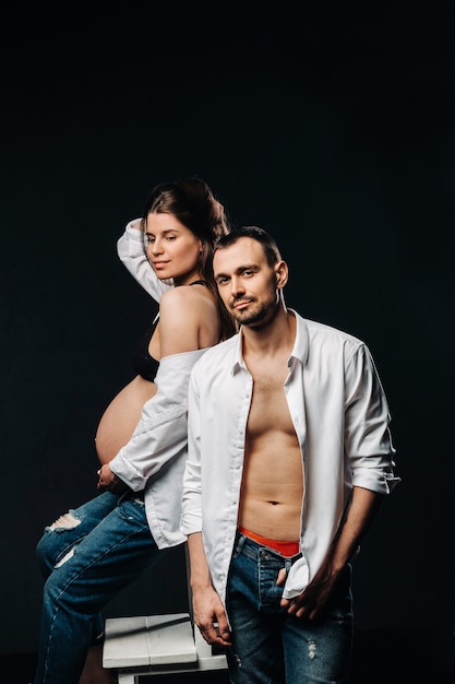 Une femme enceinte et un homme en chemise blanche et jeans dans un studio sur fond noir.