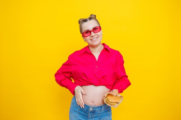 Femme enceinte joyeuse en chemise rose tenant un hamburger sur fond jaune isolé avec une expression faciale surprise et choquée Nourriture nocive pendant la grossesse Une femme enceinte mange de la restauration rapide