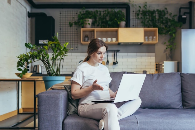 Une femme enceinte à la maison assise sur un canapé utilise un ordinateur portable pour un appel vidéo travaillant à distance