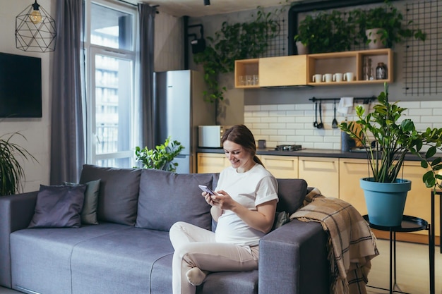 Une femme enceinte à la maison utilise le téléphone tout en étant assise sur le canapé