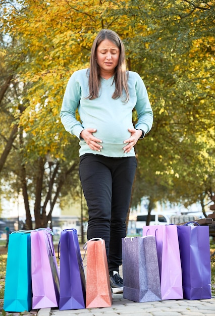 Femme enceinte malheureuse avec des sacs en papier ressentant du stress lors d'un accouchement prématuré dans le parc de la ville