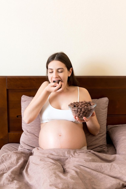 Une femme enceinte mange des boules de céréales au chocolat croquantes dans un bol reposant dans son lit Profitant de la nourriture pendant la grossesse concept