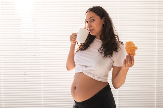 Femme enceinte mangeant un croissant et boit du café. Congé de grossesse et de maternité. Espace de copie