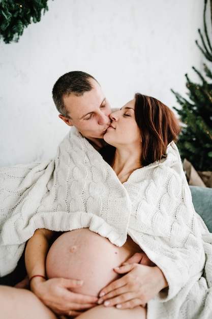 Femme enceinte et mari recouvert de plaid près de l'arbre de Noël à la maison. Joyeux Noël. Intérieur décoré. Concept de grossesse, vacances, personnes et attente.