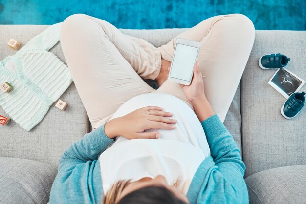 Femme enceinte et marque d'écran de téléphone et espace de maquette pour la publicité sur les réseaux sociaux et se détendre sur un canapé avec des jouets au-dessus et des vêtements pour bébé à la maison Mère smartphone 5g et lire la publication en ligne