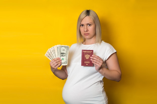 Une femme enceinte mécontente détenant un passeport avec de l'argent. Avantages pour les femmes enceintes, sur mur jaune
