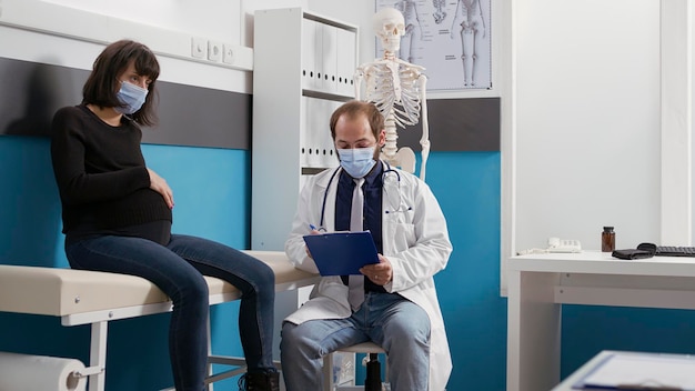 Femme enceinte et médecin avec masque facial faisant un examen, parlant de l'accouchement. Medic prenant des notes lors d'une visite de contrôle médical avec un patient qui attend un enfant dans un bureau clinique.