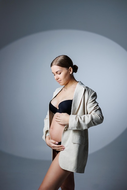 Une femme enceinte mince dans une longue veste et des sous-vêtements se tient dans le studio sur un fond blanc tenant sa main sur son ventre Sous-vêtements féminins pour femmes enceintes