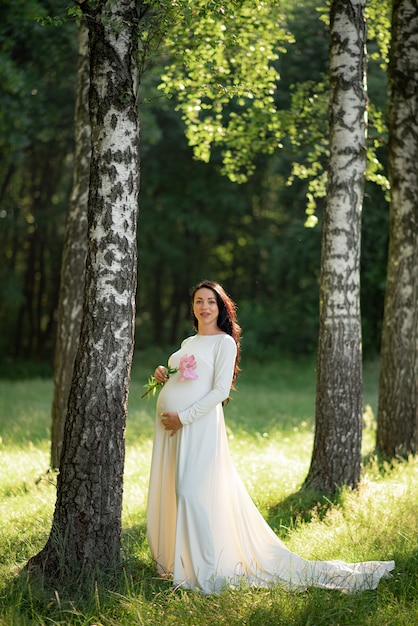 Femme enceinte posant dans une robe blanche sur fond d'arbres verts