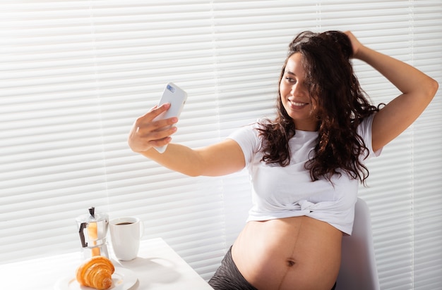 Une femme enceinte prend un selfie sur son smartphone pendant le petit-déjeuner. Concept de congé de maternité et de technologies.