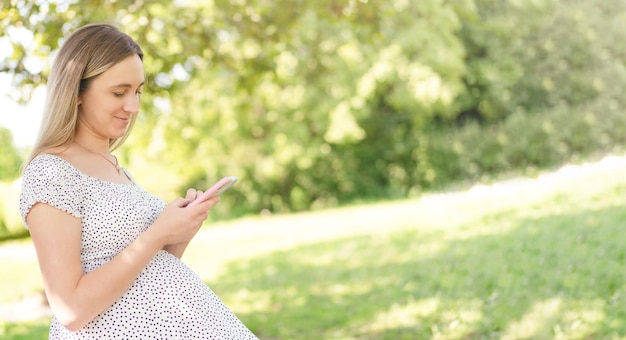 Une femme enceinte regarde son smartphone assise sur un banc dans le parc