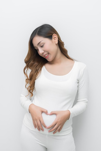 Femme enceinte en robe blanche fait la main en forme de coeur sur son ventre sur fond blanc femme asiatique