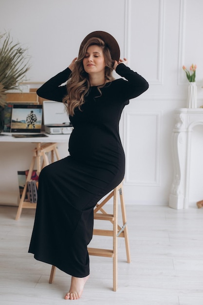 Femme enceinte en robe noire assise sur la chaise dans le studio avec un fond blanc