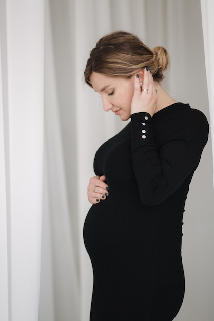 Une femme enceinte en robe noire traverse les rideaux blancs