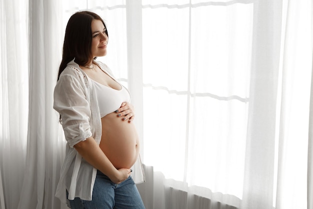 Une femme enceinte se tient près de la fenêtre dans l'appartement caressant le ventre.