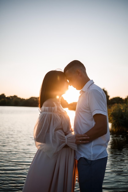 Une femme enceinte et son mari se tiennent sur la jetée et se regardent une belle rivière et le coucher du soleil les futurs parents passent du temps à l'extérieur une belle jeune mère