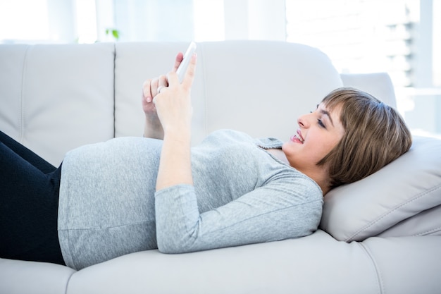 Femme enceinte souriante à l&#39;aide de smartphone en position couchée sur le canapé