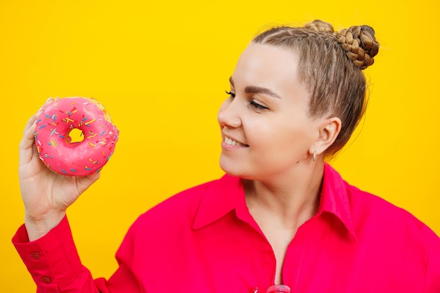 Femme enceinte souriante en chemise rose tenant de délicieux beignets sucrés sur fond jaune isolé Nourriture calorique sucrée pendant la grossesse Une femme enceinte mange des pâtisseries grasses