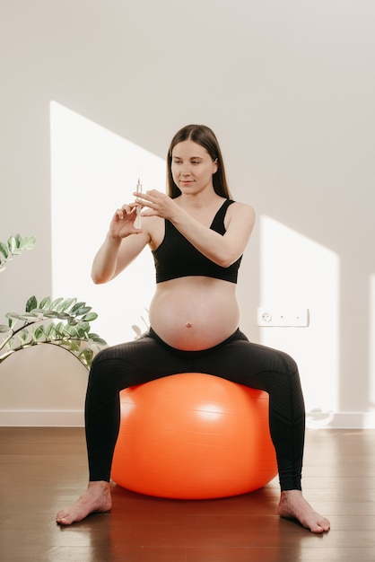 Une femme enceinte souriante est assise sur un ballon d'exercice orange et vérifie une seringue contenant des vitamines à la maison. Grossesse en bonne santé.