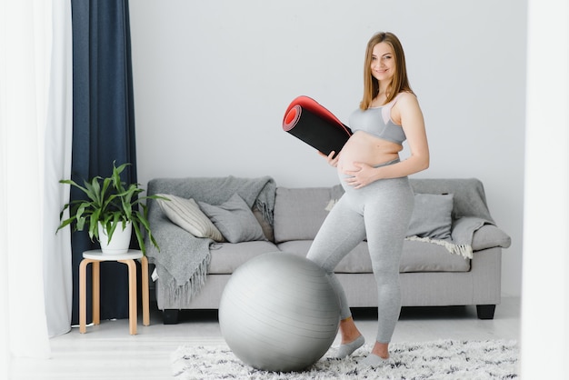 Femme enceinte souriante en tenue de sport prête pour la gymnastique matinale ou l'exercice. Une jeune femme heureuse suit un mode de vie sain pendant la grossesse, tient un tapis de yoga pour le Pilates ou s'étire.