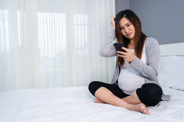 Femme enceinte stressée à l'aide d'un smartphone dans un lit