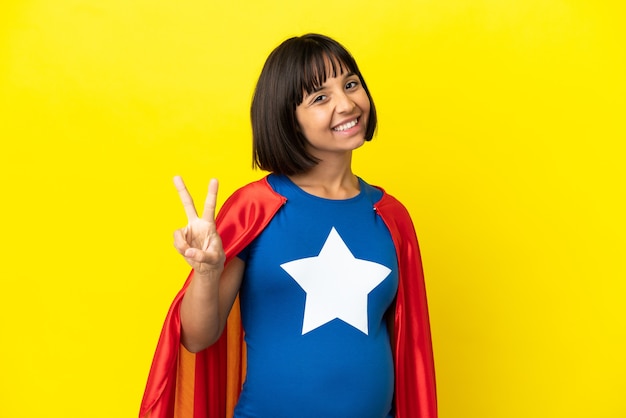 Femme enceinte de super héros isolée sur fond jaune souriant et montrant le signe de la victoire