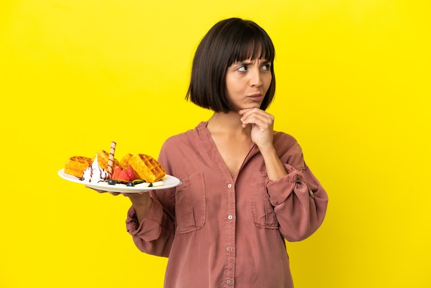 Femme enceinte tenant des gaufres isolées sur fond jaune ayant des doutes et pensant