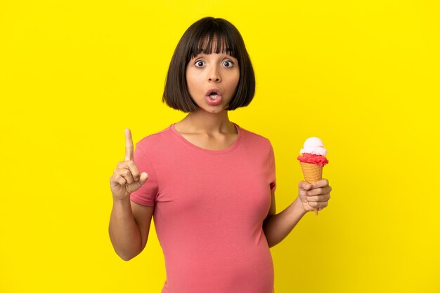 Femme enceinte tenant une glace au cornet isolée sur fond jaune dans l'intention de réaliser la solution tout en levant un doigt vers le haut