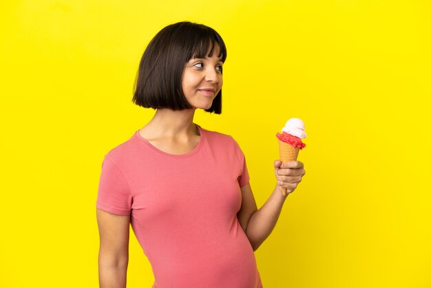 Femme enceinte tenant une glace au cornet isolée sur fond jaune regardant sur le côté et souriant