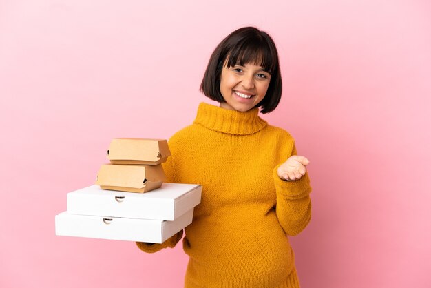 Femme enceinte tenant des pizzas et des hamburgers isolés sur fond rose se serrant la main pour conclure une bonne affaire