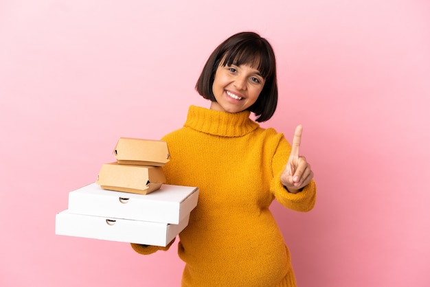 Femme enceinte tenant des pizzas et des hamburgers isolés sur une surface rose montrant et levant un doigt