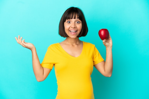 Femme enceinte tenant une pomme isolée sur fond bleu avec une expression faciale choquée