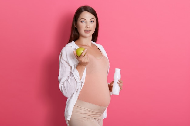 Femme enceinte tenant une pomme verte et une bouteille de lait ou de kéfir, femme enceinte posant isolé sur mur rose