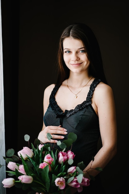 Femme enceinte tenir un bouquet de fleurs dans les mains fille embrasse un ventre rond près de la fenêtre à la maison Debout sur le côté Concept de maternité Gros plan neuf mois Douche de bébé