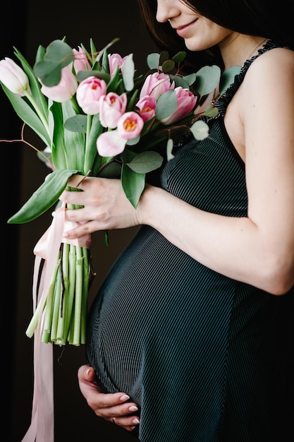 Femme enceinte tenir un bouquet de fleurs dans les mains fille embrasse un ventre rond près de la fenêtre à la maison Debout sur le côté Concept de maternité Gros plan neuf mois Douche de bébé