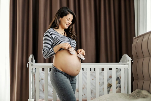 Une femme enceinte tient des chaussures de bébé tout en montrant son ventre nu avec des vergetures