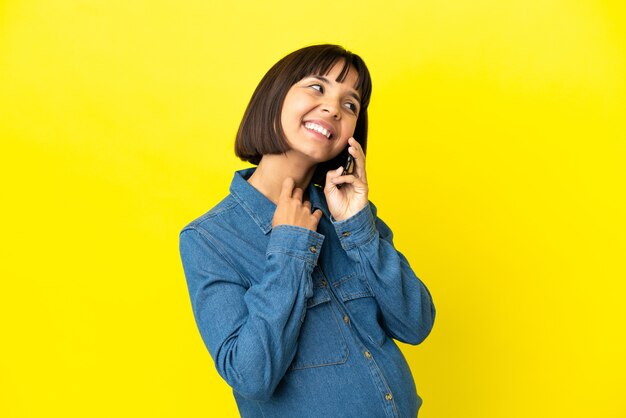 Femme enceinte utilisant un téléphone portable isolé sur fond jaune en levant tout en souriant