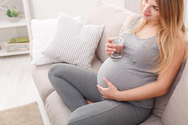 Femme enceinte avec un verre d'eau assis sur un canapé. Jeune blonde enceinte reste à la maison, copiez l'espace. Grossesse, soins de santé, concept de soif