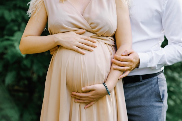 Une Femme Enceinte Vêtue D'une Longue Robe Beige Tient Ses Mains Sur Son Ventre Alors Que Son Mari Et Son Futur Papa Embrassent Sa Femme. Gros Plan Des Bras Et De L'abdomen. Femmes Enceintes